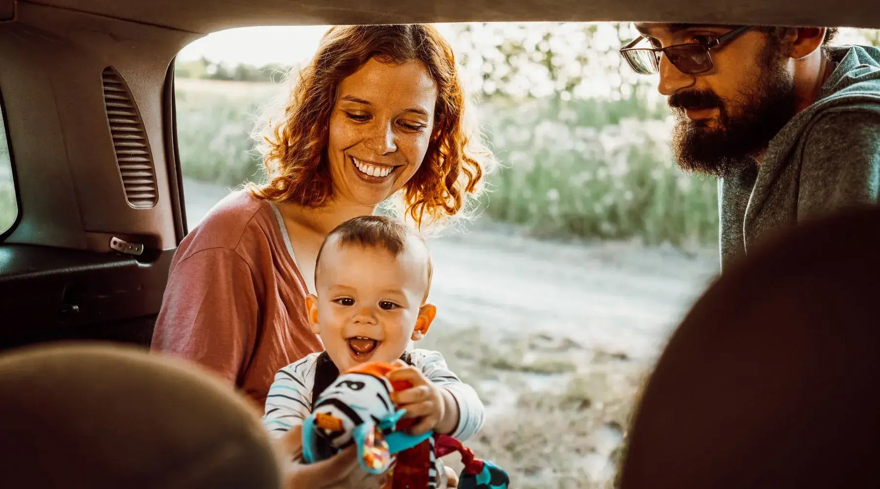 A happy family in a car with a baby holding a toy. The parents smile as they interact with their joyful child, with a scenic background visible outside the car.