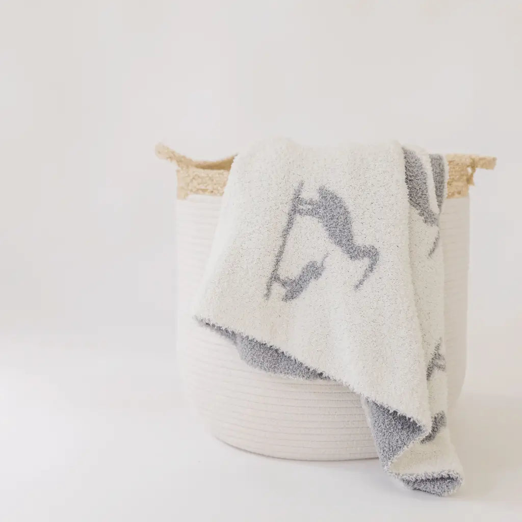 white blanket with gray herons draped over a white bell shaped rope basket with corn husk details and handlesr