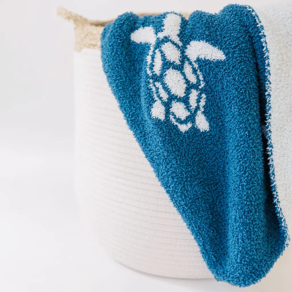 blue blanket with white sea turtles draped over a white bell shaped rope basket with corn husk details and handlesr