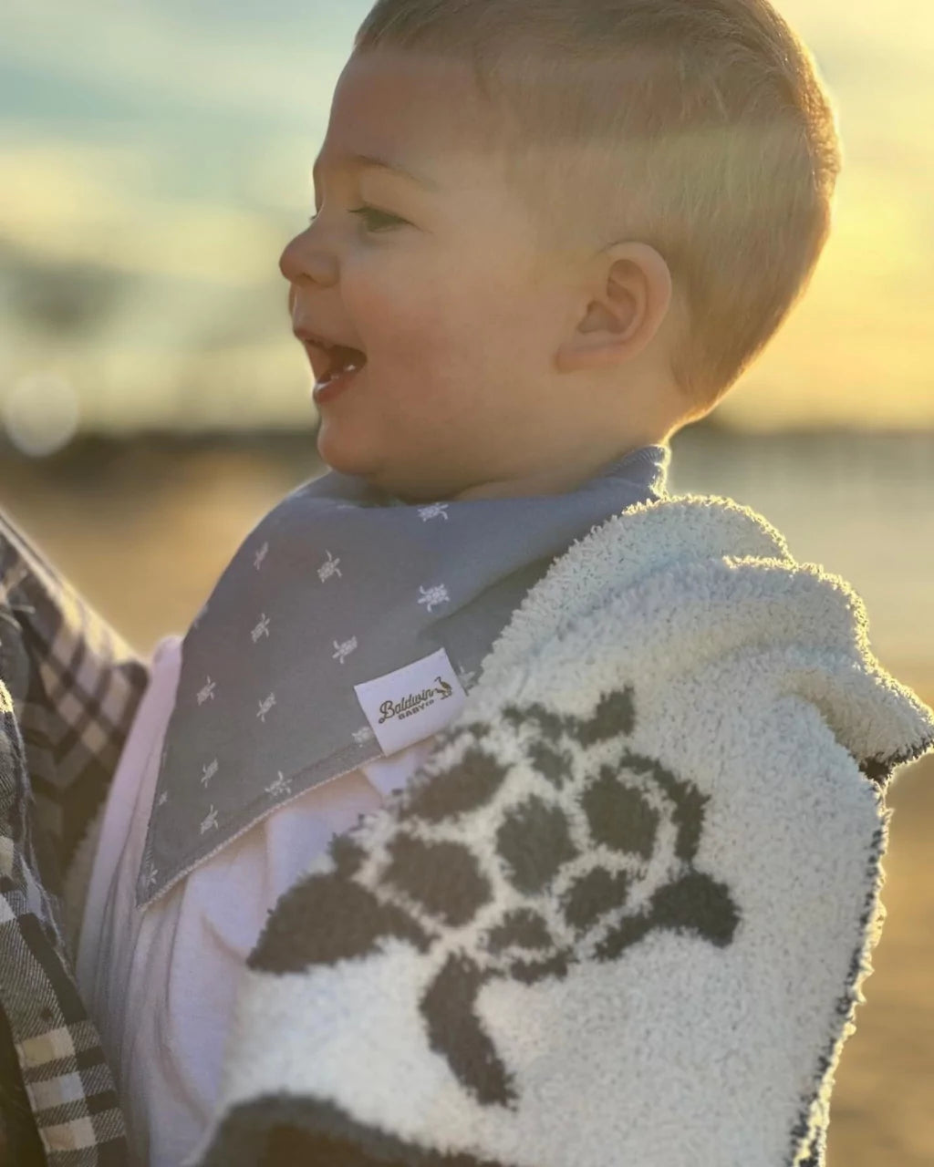 Toddler boy wearing a blue with with white sea turtle pattern bandana bib by Baldwin Baby Co wrapped in a white blanket with blue sea turtles