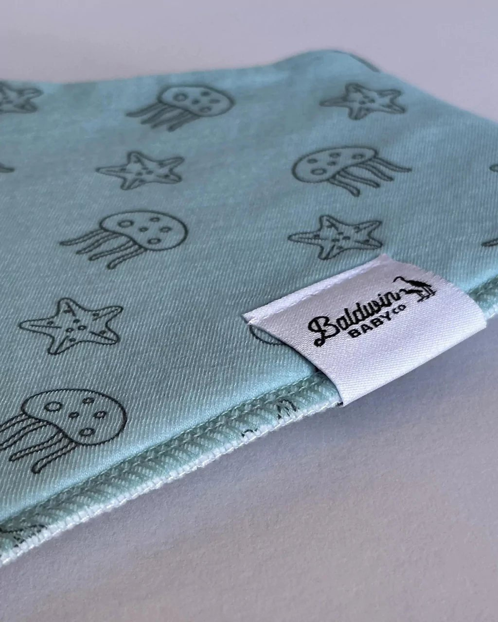 Close up of light blue with starfish and jellyfish pattern bandana bib with a white Baldwin Baby Company logo tag