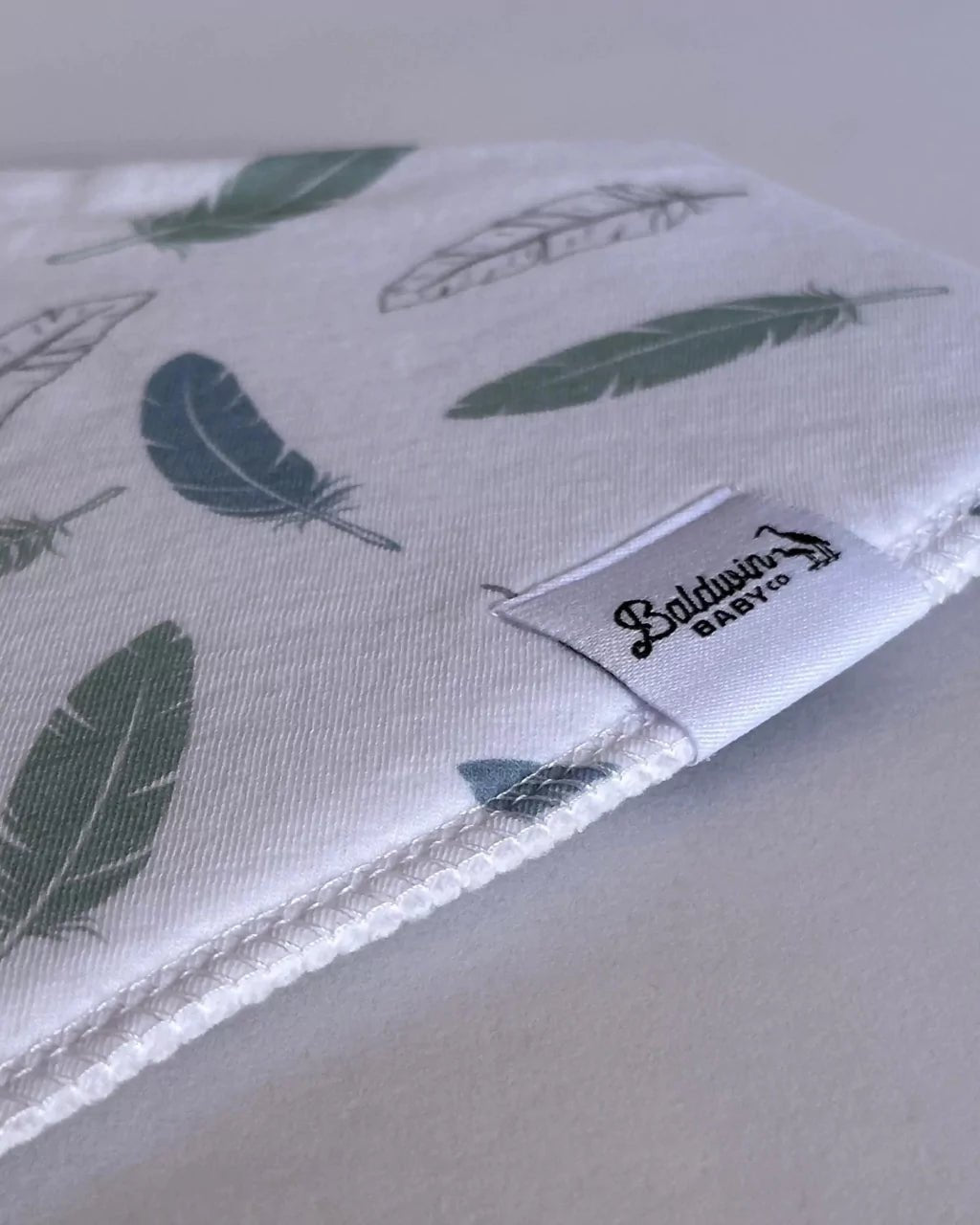 Close up of white with a green and transparent feather pattern bandana bib with a white Baldwin Baby Company logo tag