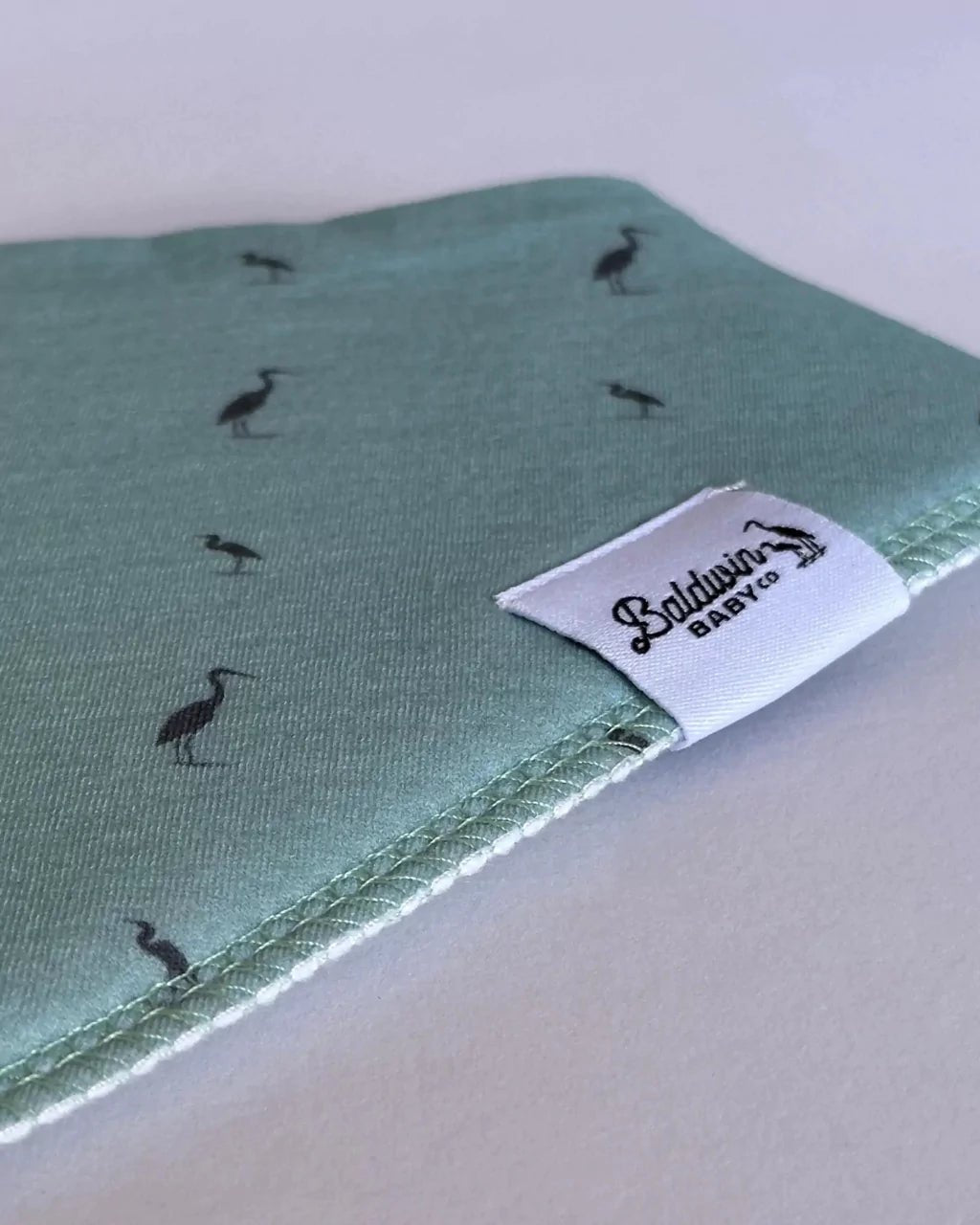 Close up of green with a dark gray heron pattern bandana bib with a white Baldwin Baby Company logo tag