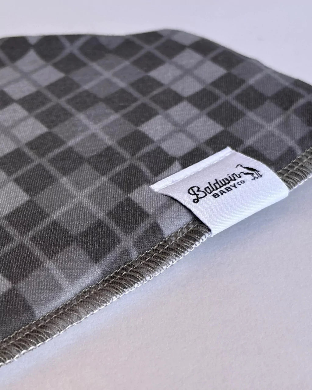 Close up of a grayscale plaid or argyle pattern bandana bib with a white Baldwin Baby Company logo tag