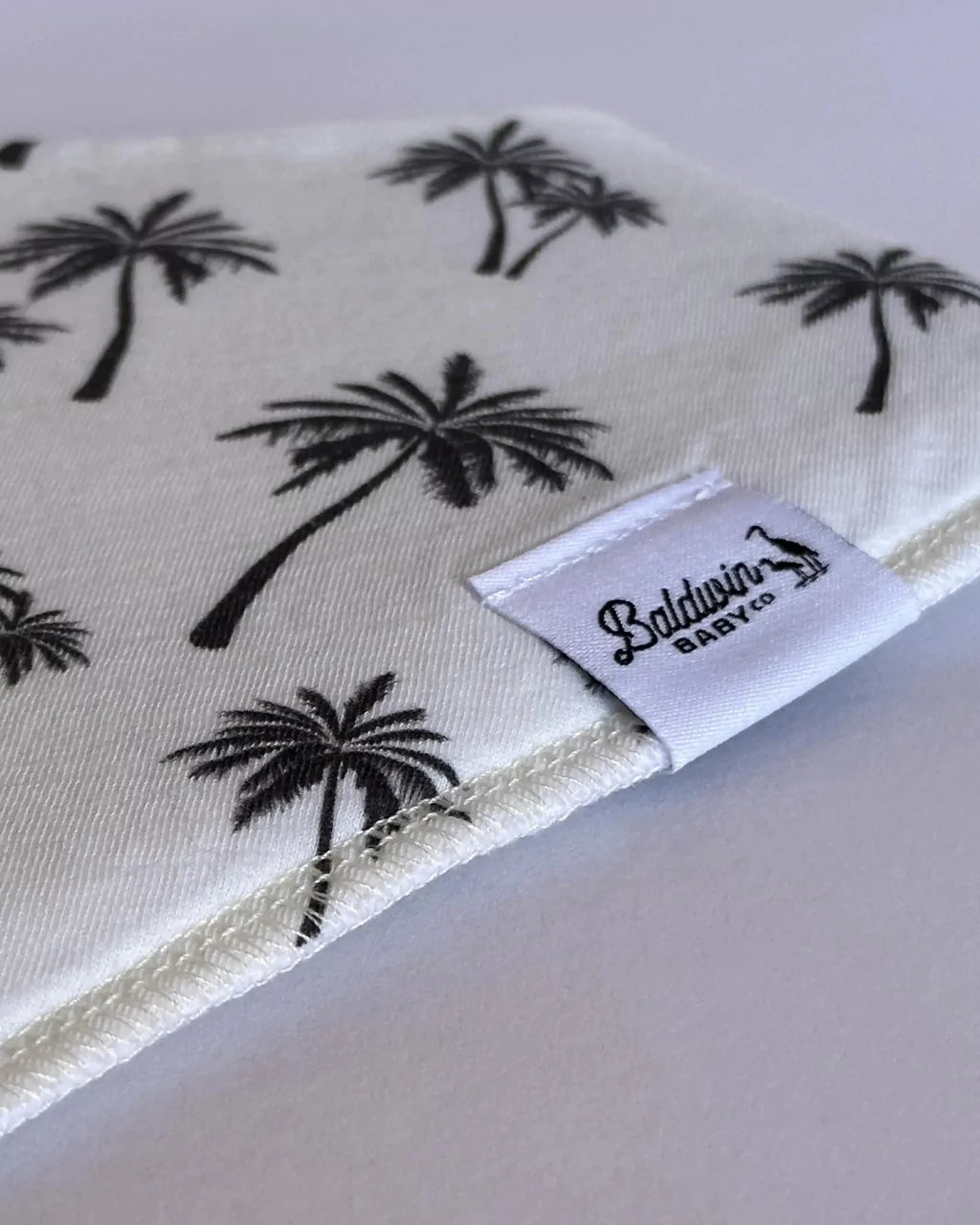 Close up of a creamy white black palm tree pattern bandana bib with a white Baldwin Baby Company logo tag