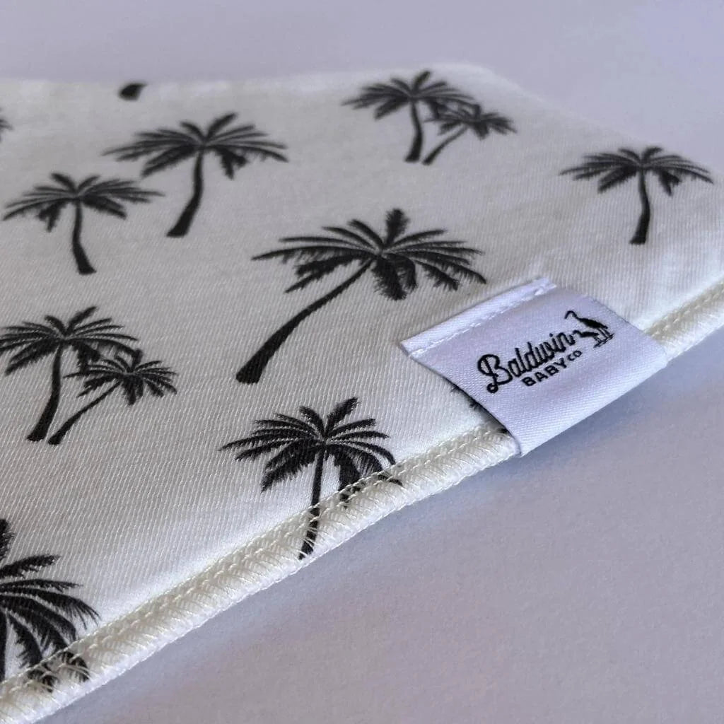 Close up of a creamy white black palm tree pattern bandana bib with a white Baldwin Baby Company logo tag
