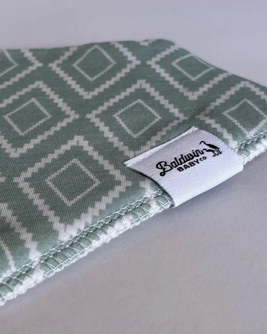 Close-up of a green with white western or Aztec style diamond pattern bandana bib with a white Baldwin Baby Company logo tag