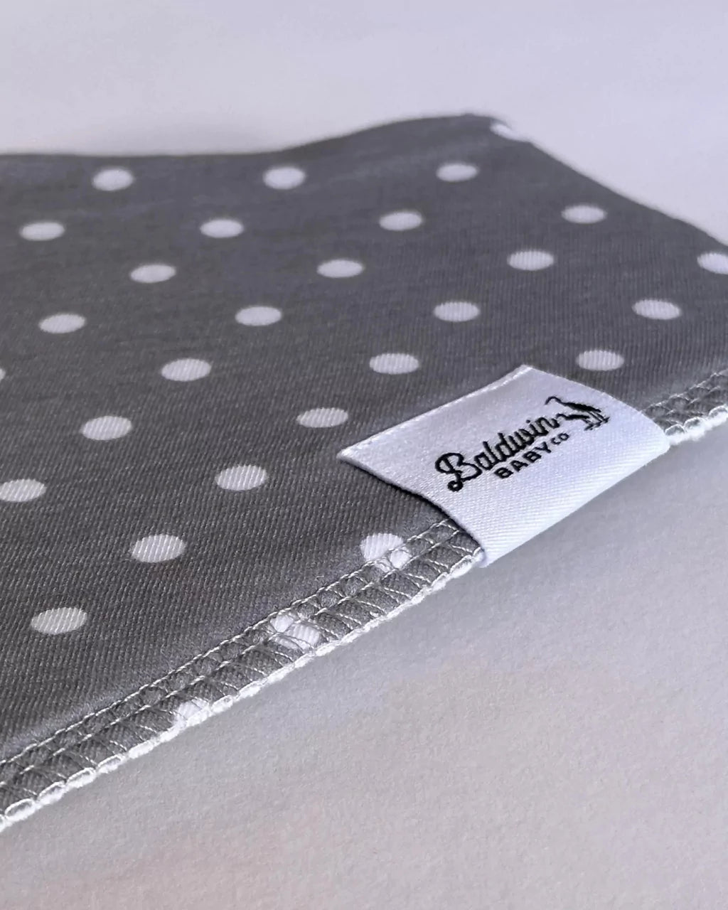 Close up of a gray with white polka dot pattern bandana bib with a white Baldwin Baby Company logo tag