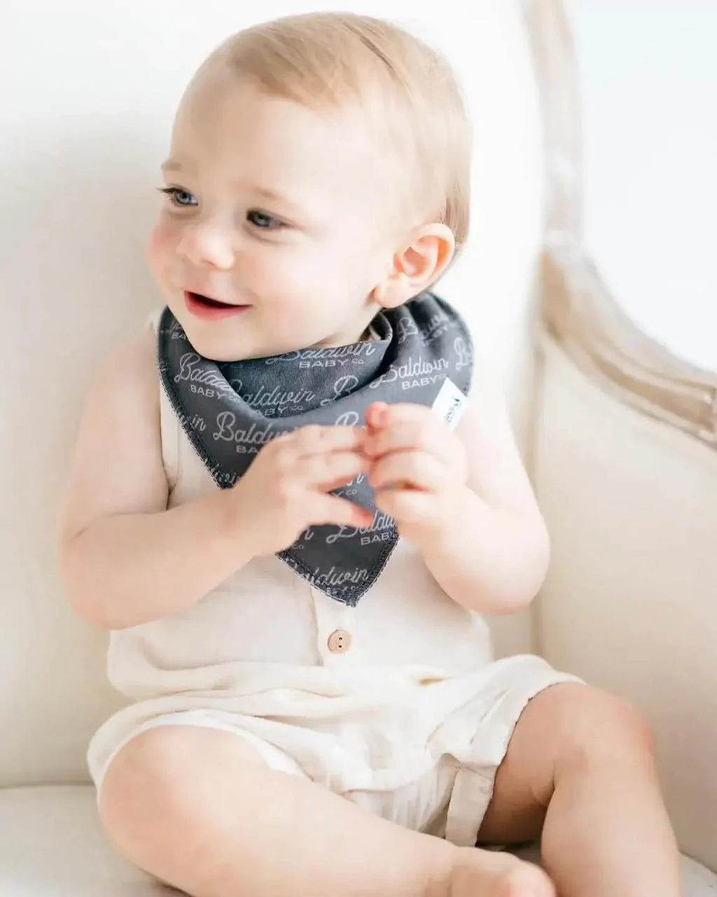 Little boy sitting in white chair while wearing a charcoal with a light gray Baldwin Baby Co pattern bandana bib