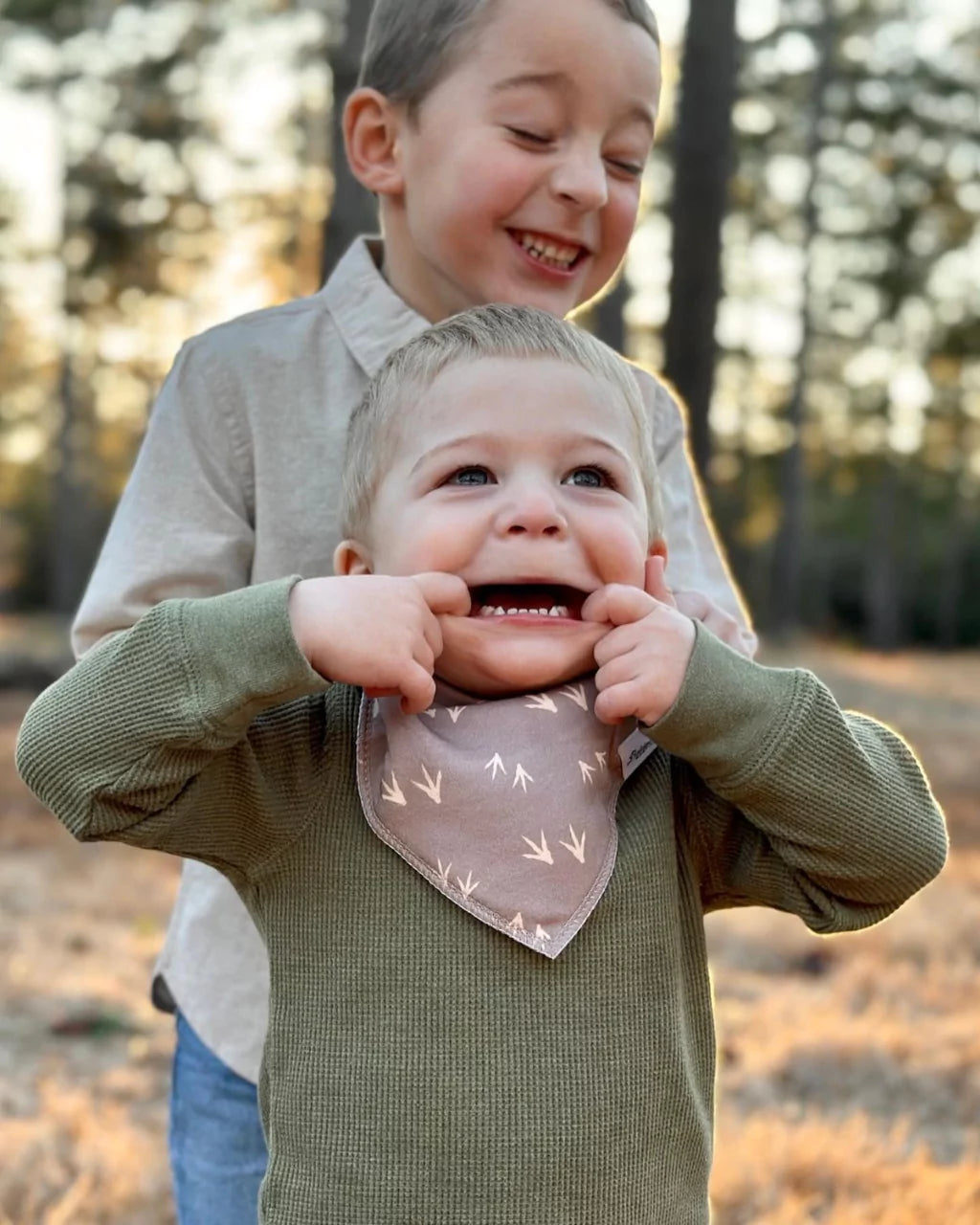 Toddler boy making a silly face wearing a brown with tan turkey track pattern bandana bib by Baldwin Baby Co standing infront of older boy 