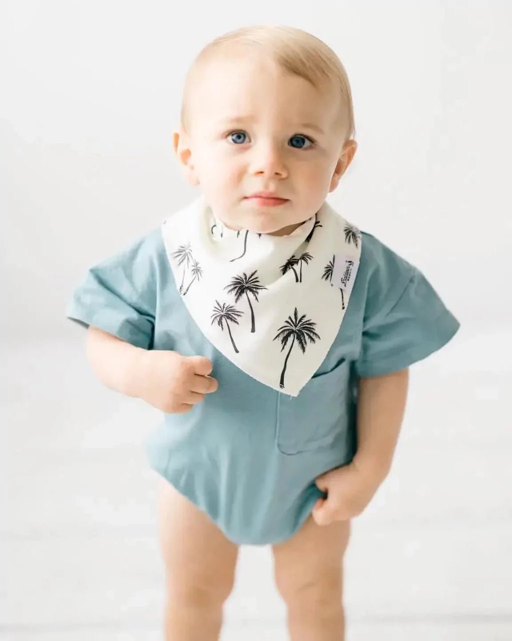 Little boy standing on white background while wearing a white and black palm tree pattern bandana bib