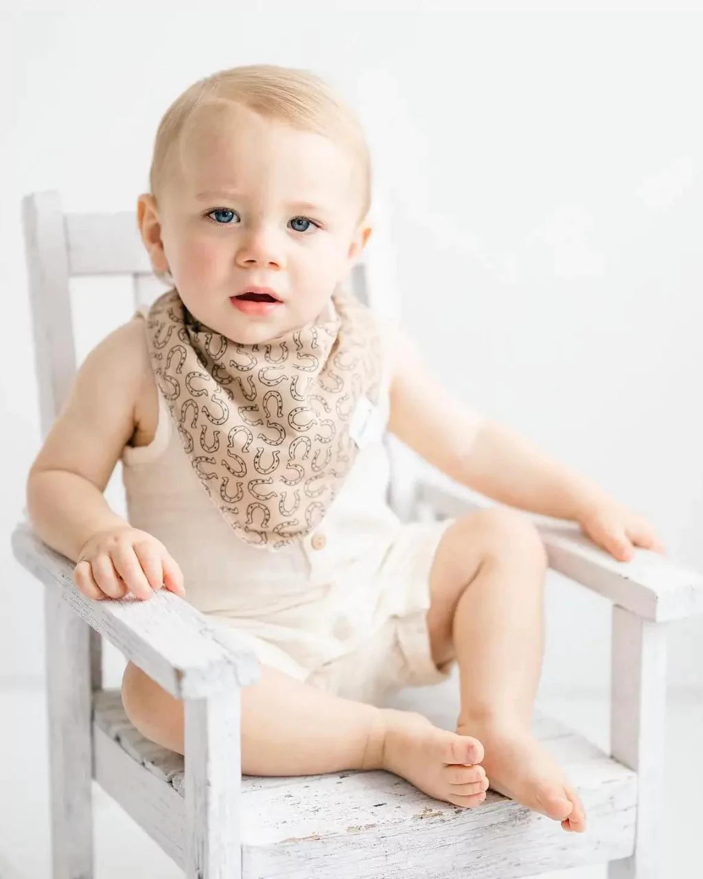 Little boy sitting in a rocking chair while wearing a tan with brown horseshow pattern bandana bib