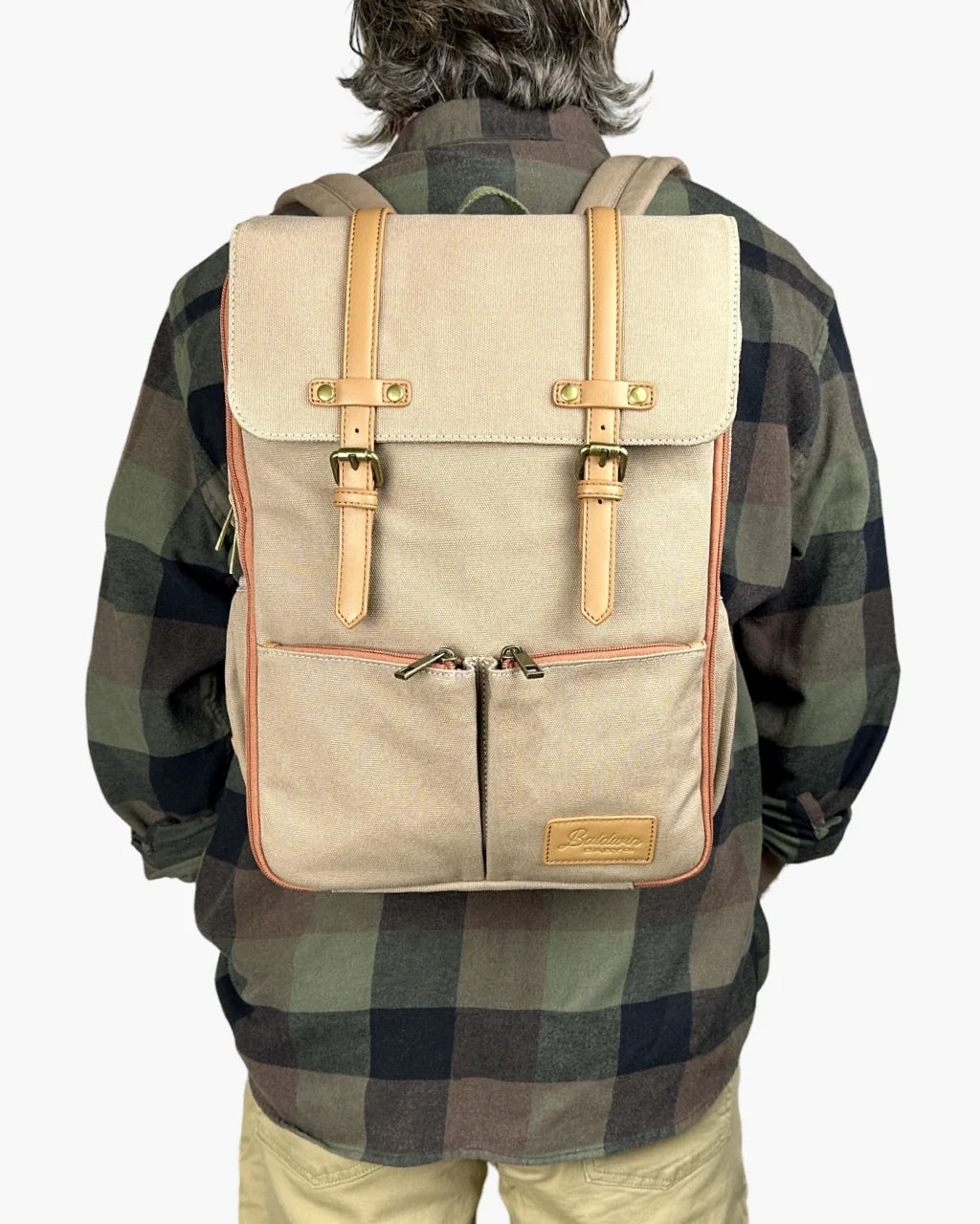 A man wearing a beige canvas diaper backpack with tan leather straps and brass buckles, featuring front zippered pockets and a rectangular silhouette