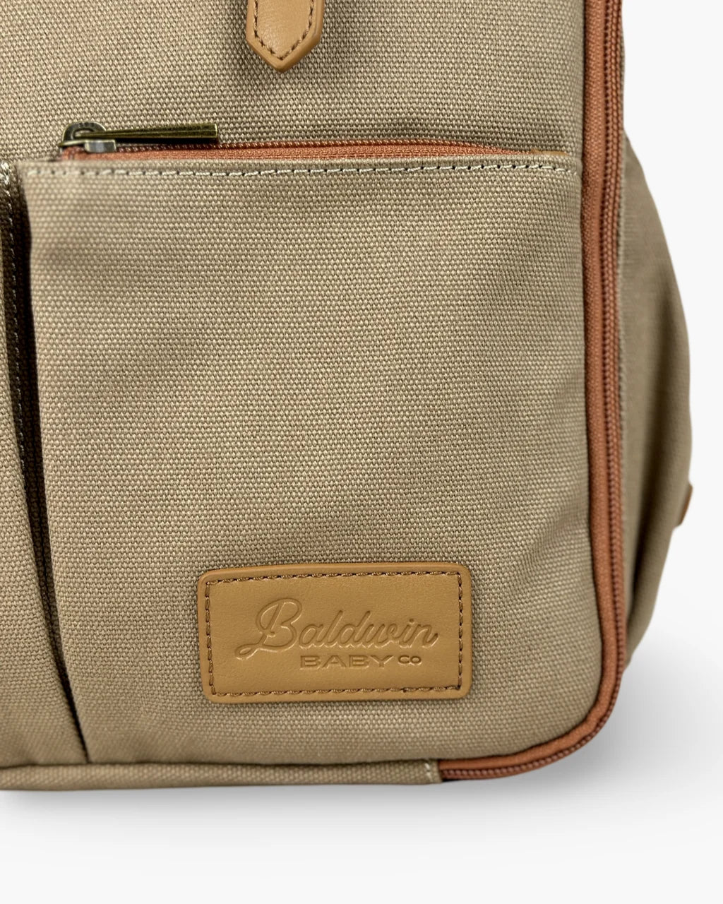 Close-up of the Baldwin Baby Co. logo embossed on a tan leather patch at the bottom of the beige canvas diaper backpack.