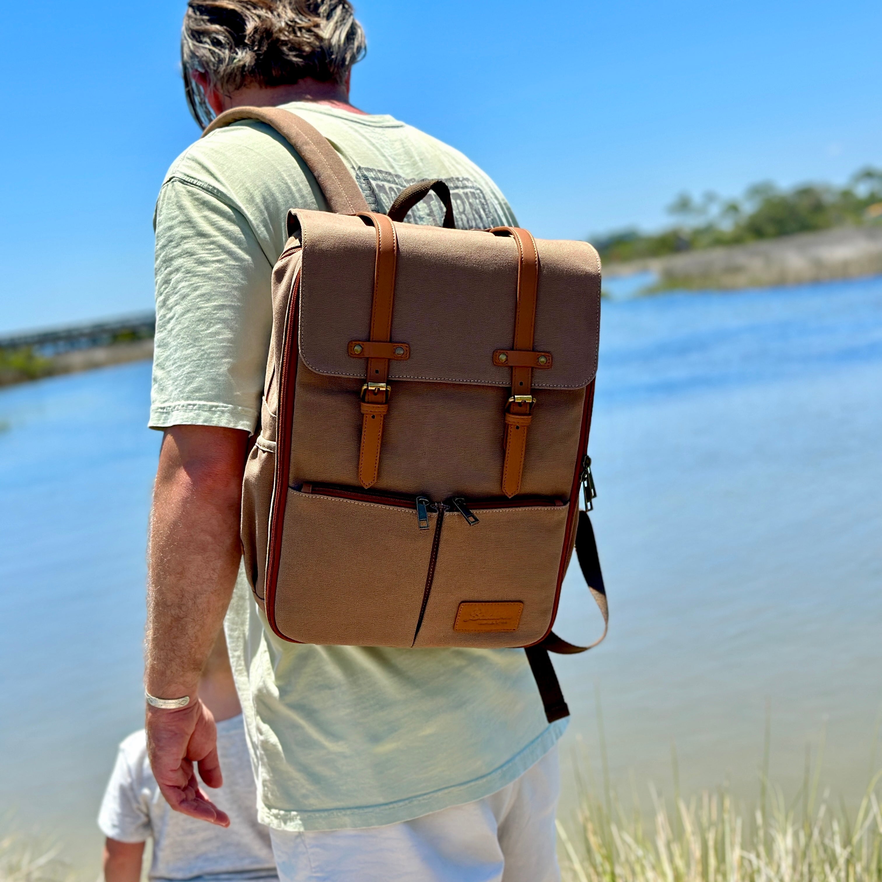 man wearing canvas diaper bag backpack while watching toddler overlooking the water