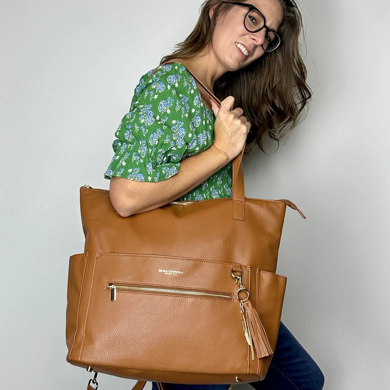 The image shows a person wearing a green floral blouse and jeans, holding the Cognac Lillian Diaper Bag. The bag is styled with a gold zipper and a tassel charm, showcasing its spacious and fashionable design.