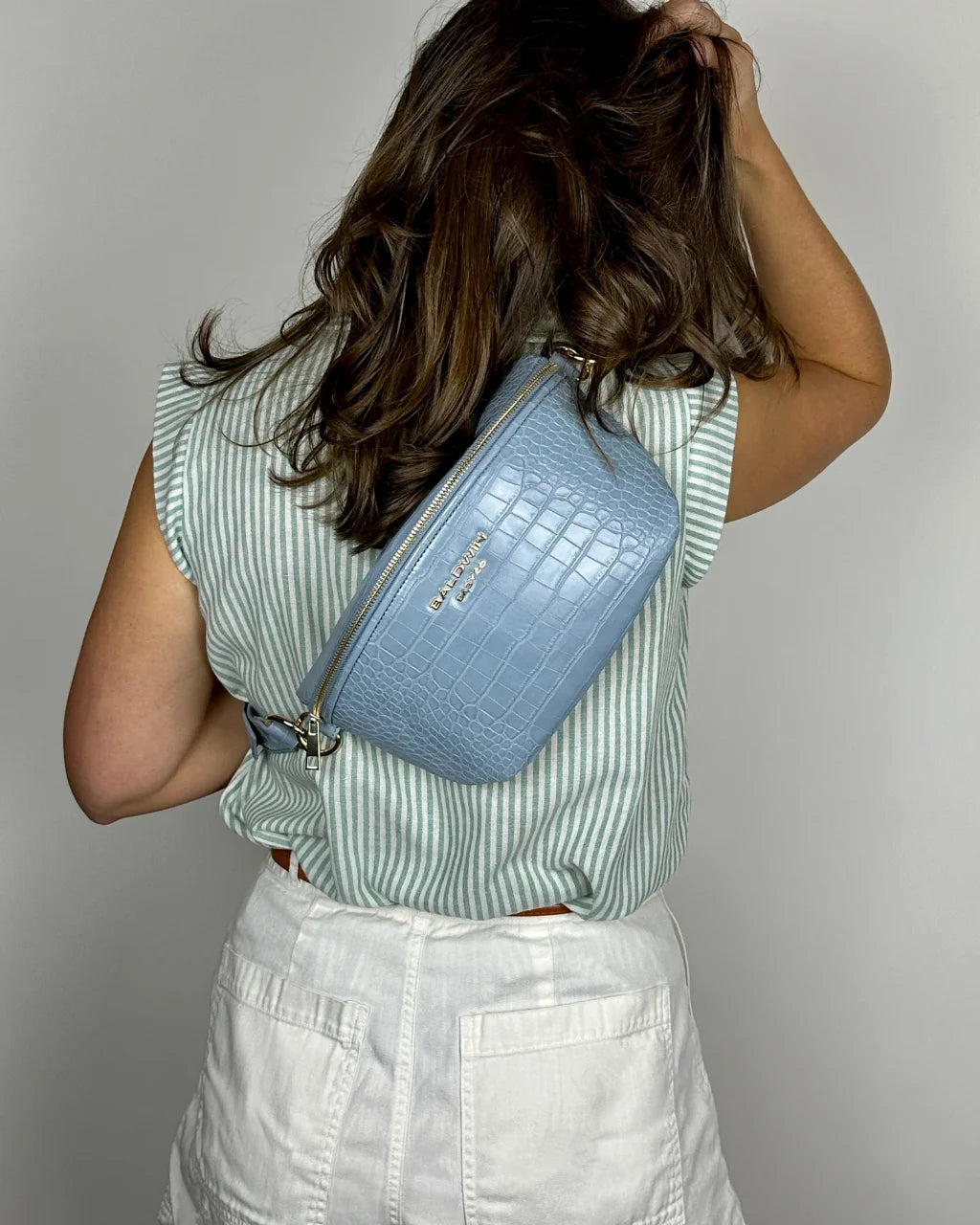 Women wearing green and white shirt and white shorts wearing a blue faux crocodile crossbody belt bag on her back, showcasing its stylish and adjustable strap with gold hardware