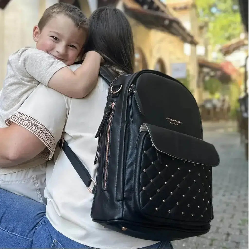 Boy smiling and hugging a kneeling woman wearing a black leather backpack diaper bag with rose gold studs quilted on the front pocket 
