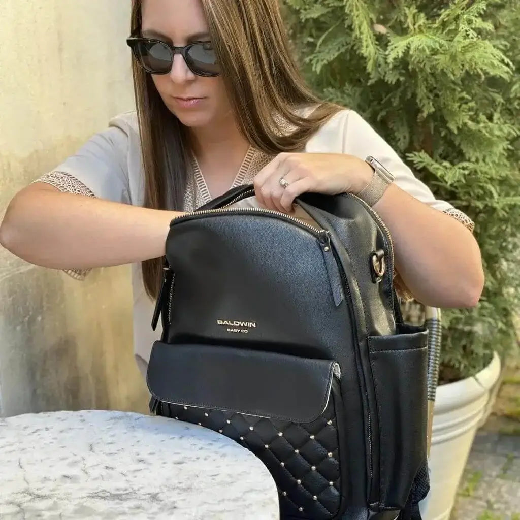 Women wearing sunglasses reaching into black leather backpack diaper bag with rose gold studs quilted on front pocket 