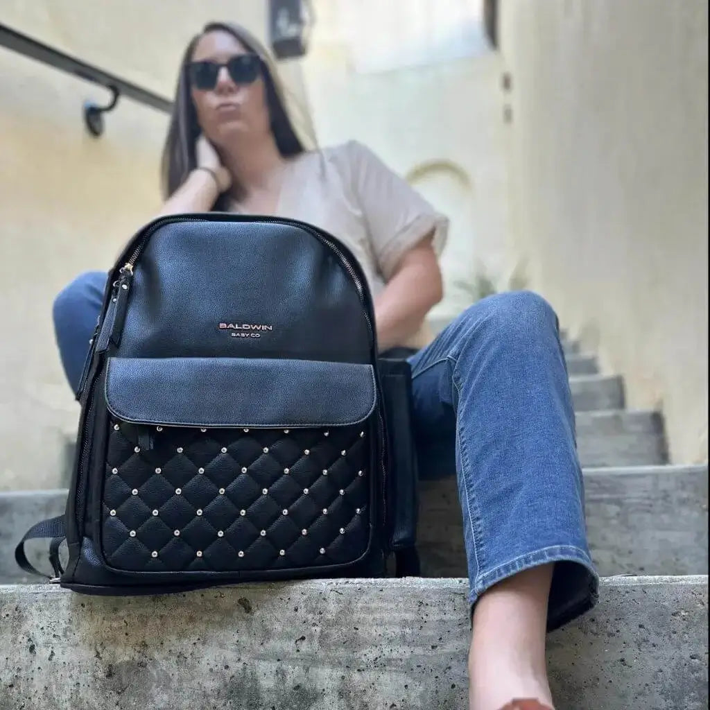Black leather backpack diaper bag with rose gold studs quilted on front pocket in front of women sitting on stairs 