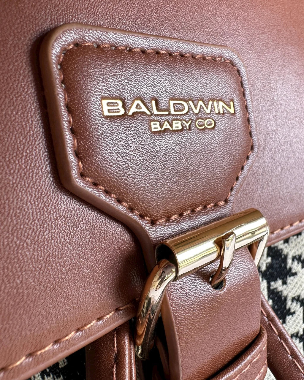 Close-up view of the Baldwin Baby Co logo embossed in gold on a brown vegan leather patch of the houndstooth diaper bag backpack.