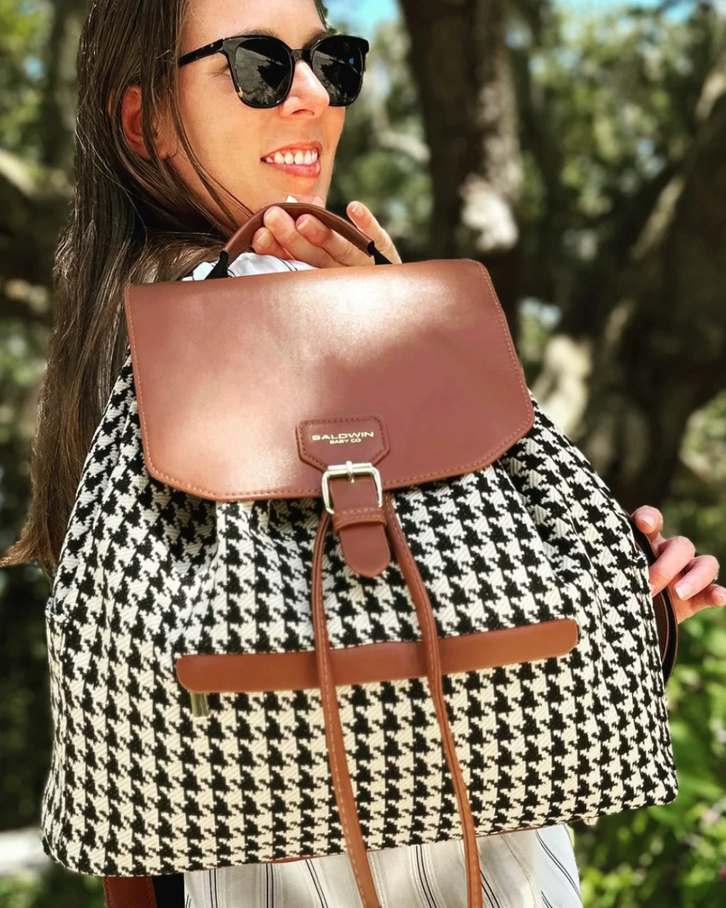 Woman outdoors carrying a houndstooth diaper bag backpack with brown vegan leather details and adjustable straps, side view.