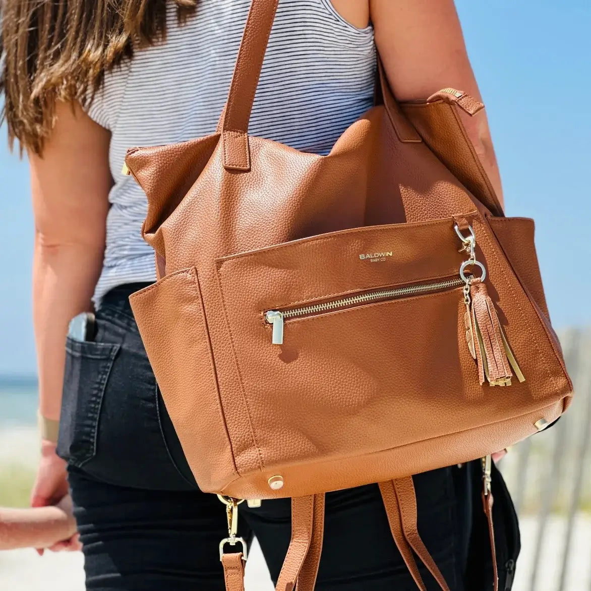 Woman on the beach wearing brown leather diaper bag with leather and gold feather tassel by the shoulder straps