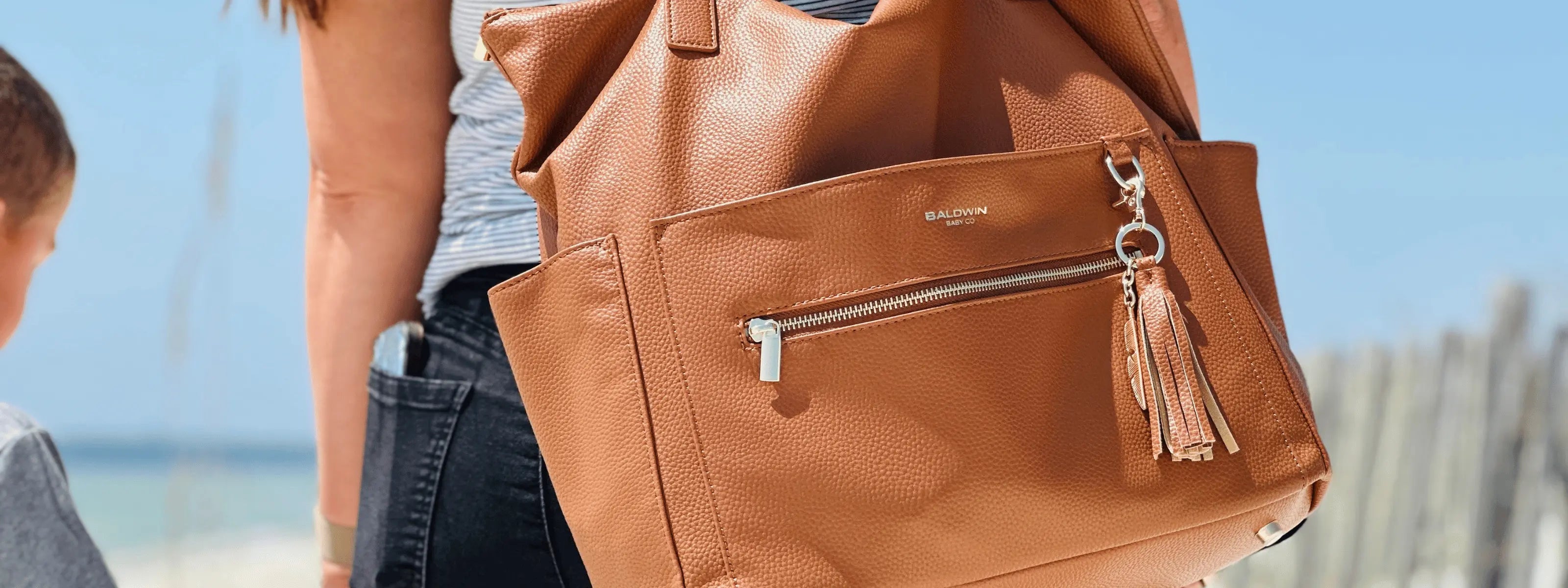 Woman holding hands with children on the beach wearing brown leather diaper bag with leather and gold feather tassel by the shoulder straps