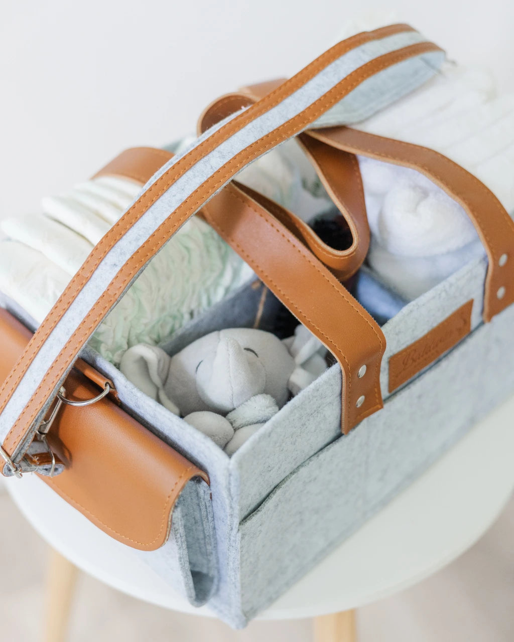 Gray felt diaper caddy with brown leather handles, filled with baby items, on a wooden table.