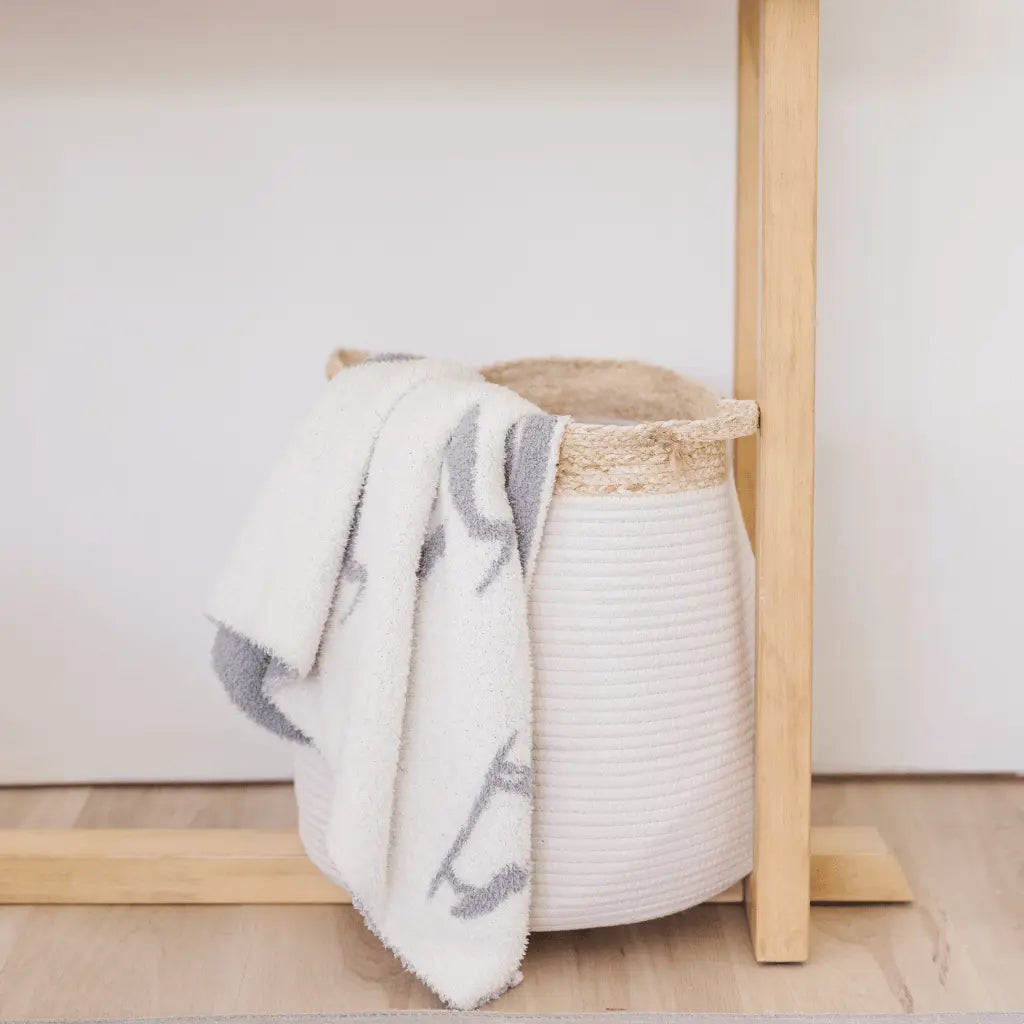 white blanket with gray herons draped over a white bell shaped rope basket with corn husk details and handlesr