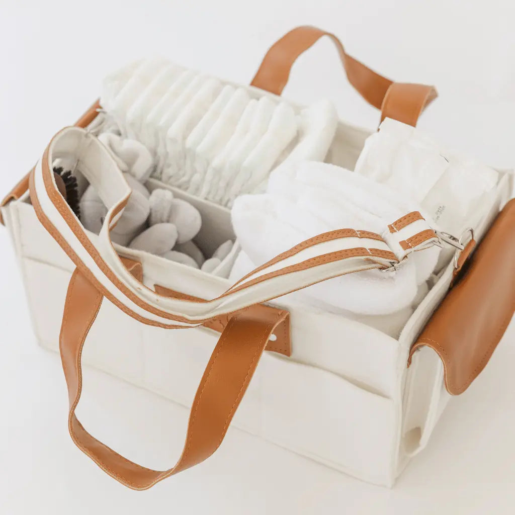 White felt diaper caddy with brown leather handles, filled with baby items, on a wooden table.