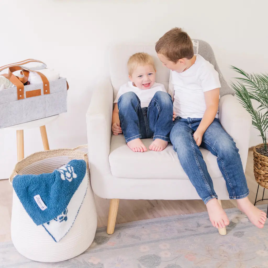 two young boys sitting in a chair with a white bell shaped rope basket with corn husk details and handles and a blue blanket drapped over the side