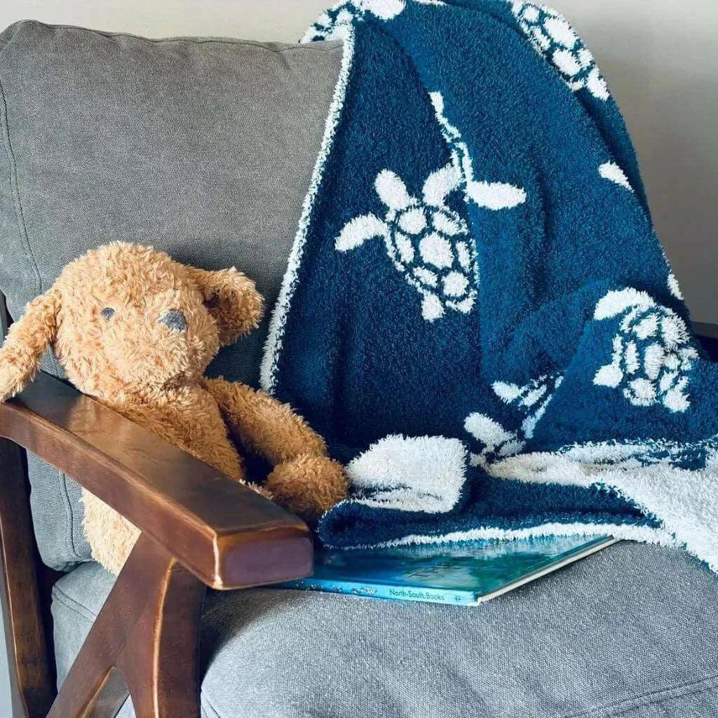 Blue with white sea turtle pattern blanket draped over a gray chair with teddy bear and book