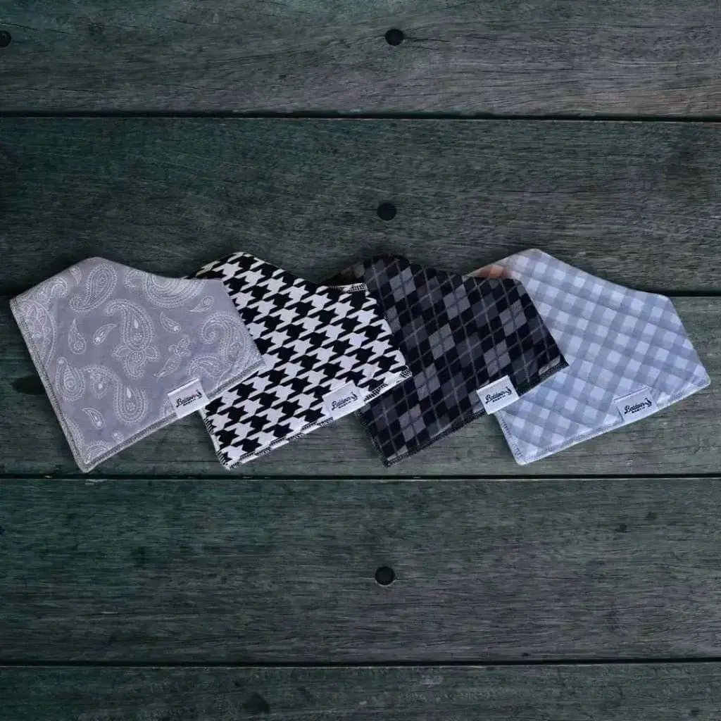 Set of four bandana bibs on a wood background. Gray with white paisley, traditional white and black houndstooth, grayscale plaid or argyle, and timeless gray and white checkered patterns