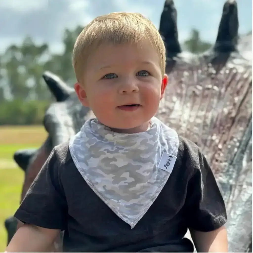 Little boy sitting on dinosaur statue while wearing a gray and white camo pattern bandana bib