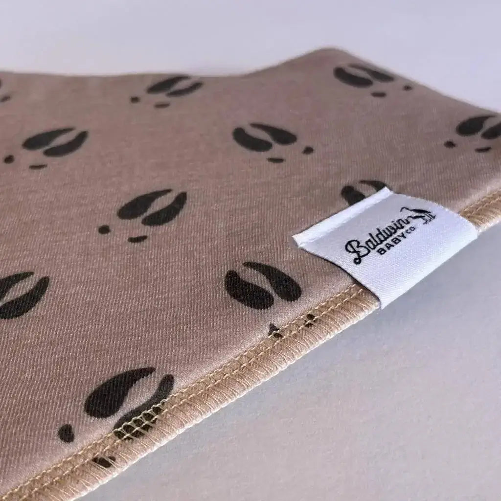 Close up of tan with black deer track pattern bandana bib with a white Baldwin Baby Company logo tag