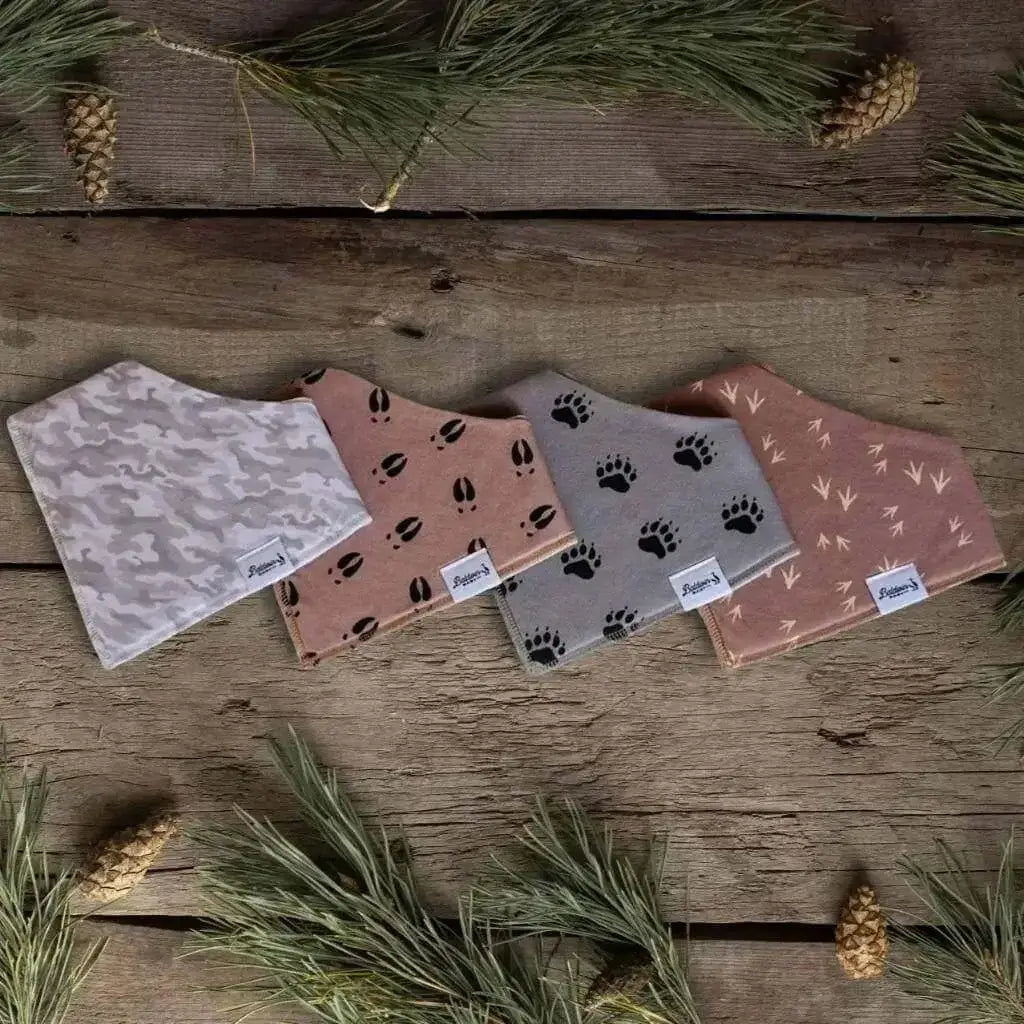 Set of four bandana bibs on a wood background with pine needles and pinecones. Gray and white camo, tan with black deer tracks, gray with black bear paw tracks, and brown with tan turkey track patterns