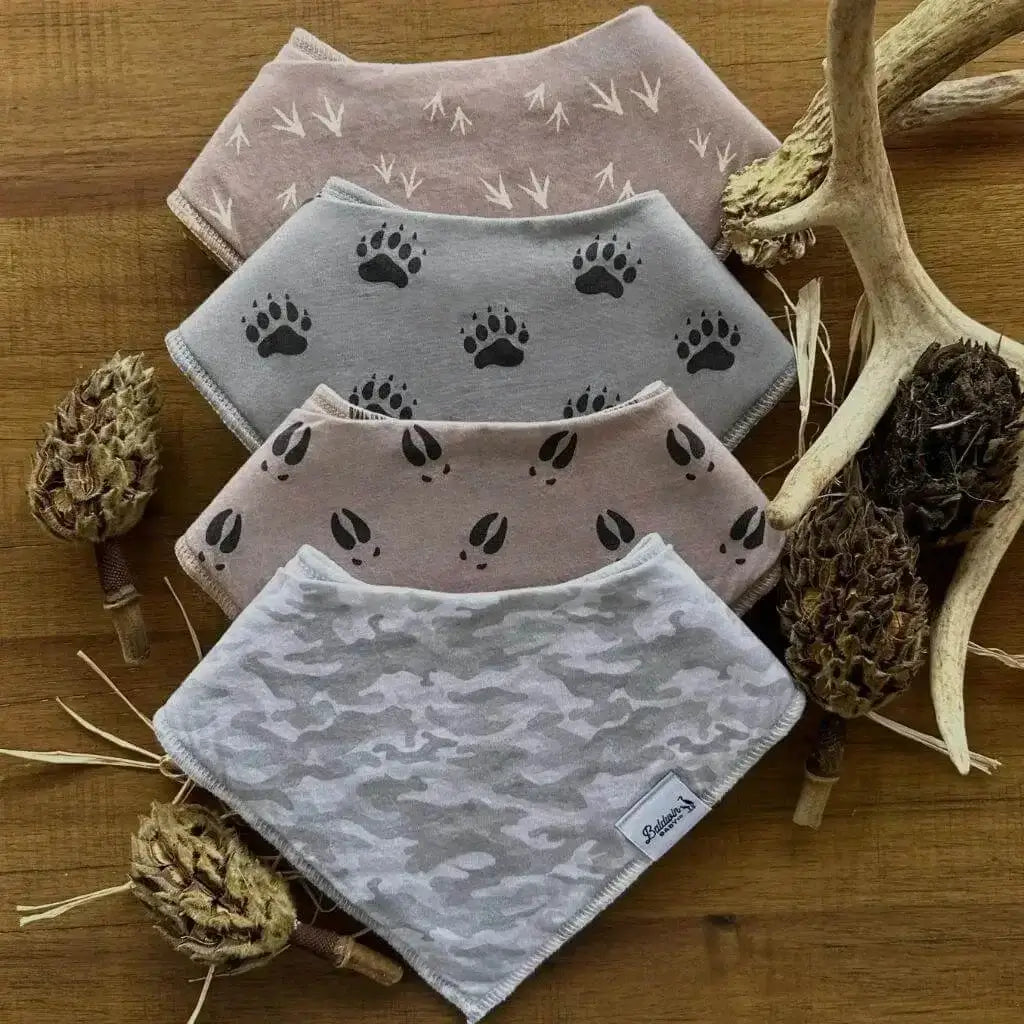 Set of four bandana bibs on a wood background and deer antlers. Gray and white camo, tan with black deer tracks, gray with black bear paw tracks, and brown with tan turkey track patterns