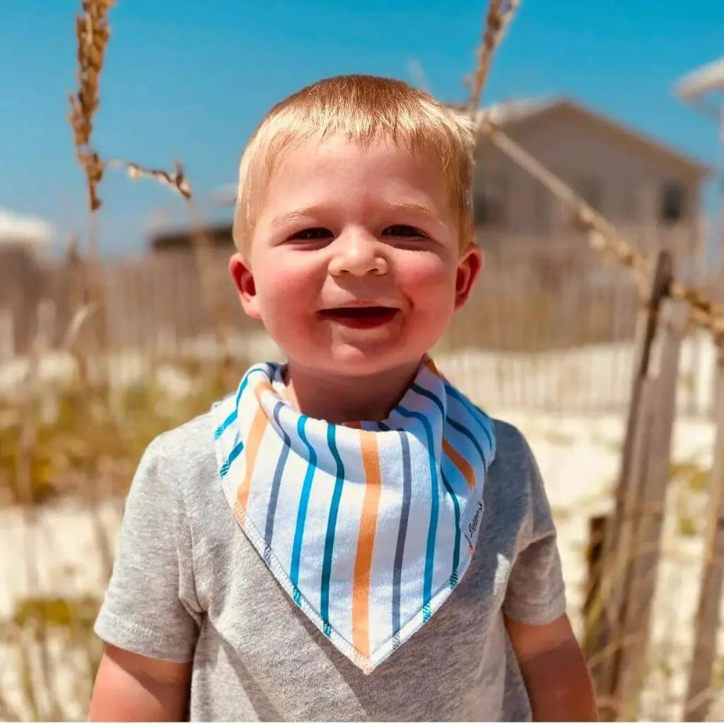 Little boy on the beach while wearing a white, blue, gray, and orange striped pattern bandana bib