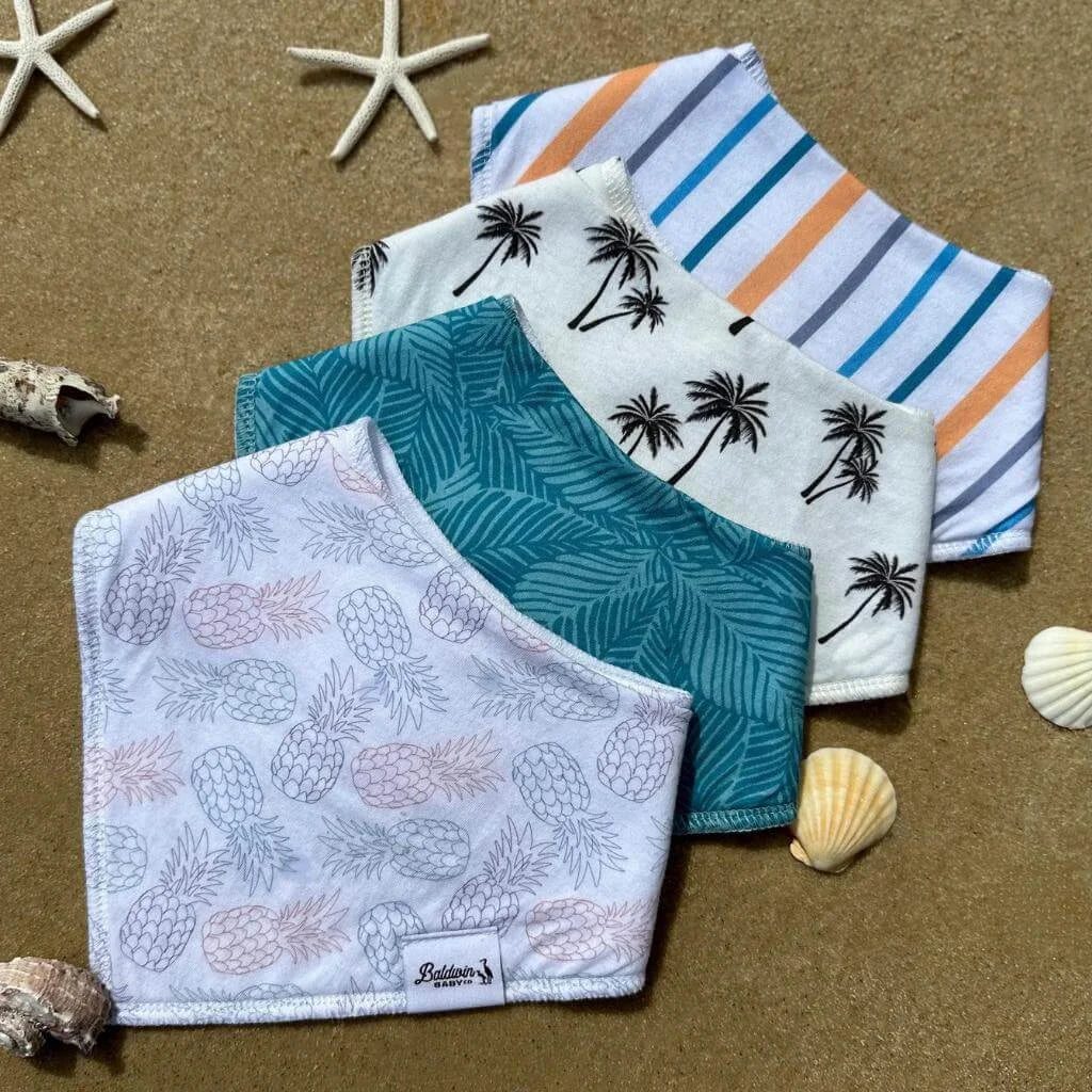 Set of four bandana bibs on sand with seashells and starfish in the background. White, blue, gray, and orange stripes; white with blue, pink, and green pineapples; green monstera; and creamy white black palm tree patterns