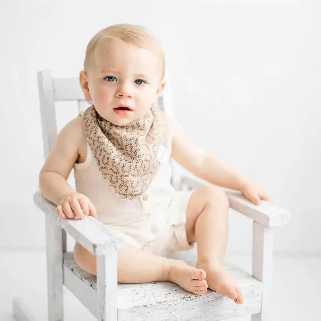 Little boy sitting in a rocking chair while wearing a tan with brown horseshow pattern bandana bib