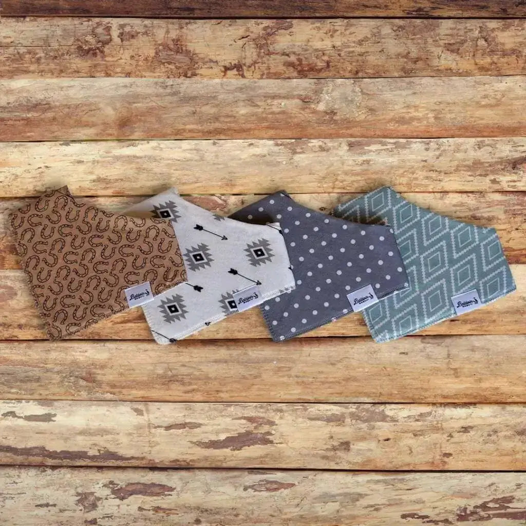 Set of four bandana bibs on a wood background. Tan with brown horseshoes, creamy white and black arrows, gray with white polka dots, and green with white western or Aztec-style diamond patterns